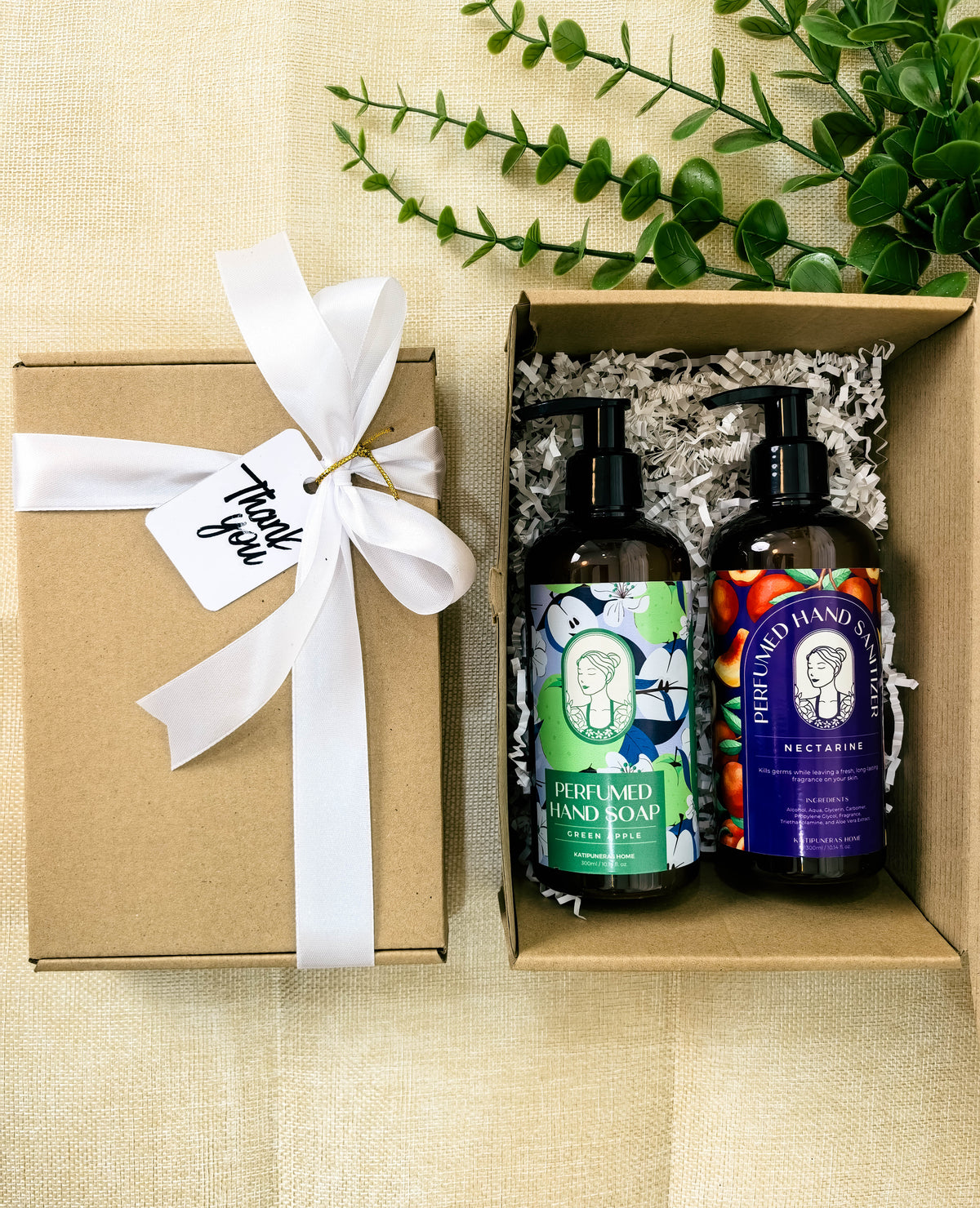 A gift set containing two pump bottles of hand wash and hand sanitizer in a kraft box with shredded paper, next to a closed kraft box tied with a white ribbon and tag, on a beige fabric background with green leaves.
