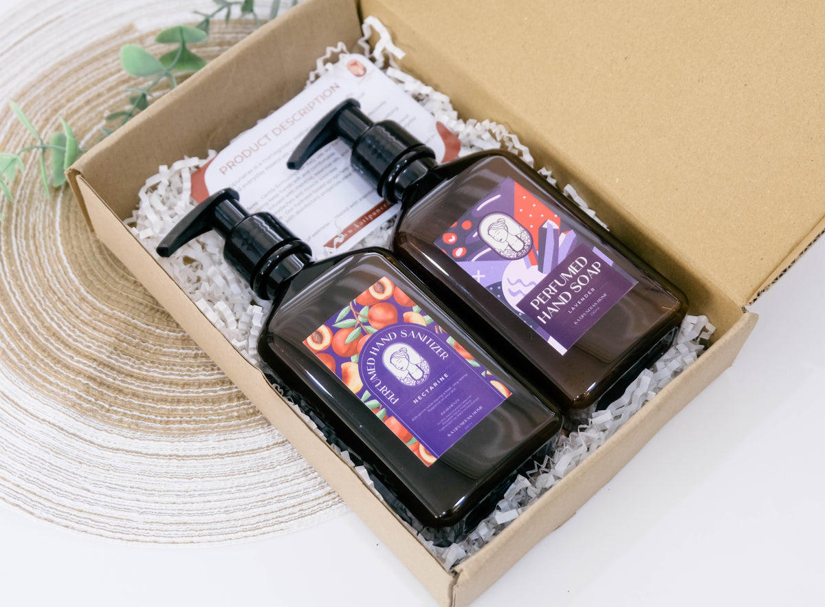 A gift box containing two dark amber bottles of hand soap and hand sanitizer with black pumps, nestled in white crinkle paper. One bottle is labeled 'Lavender' and the other 'Jo Malone Nectarine.'