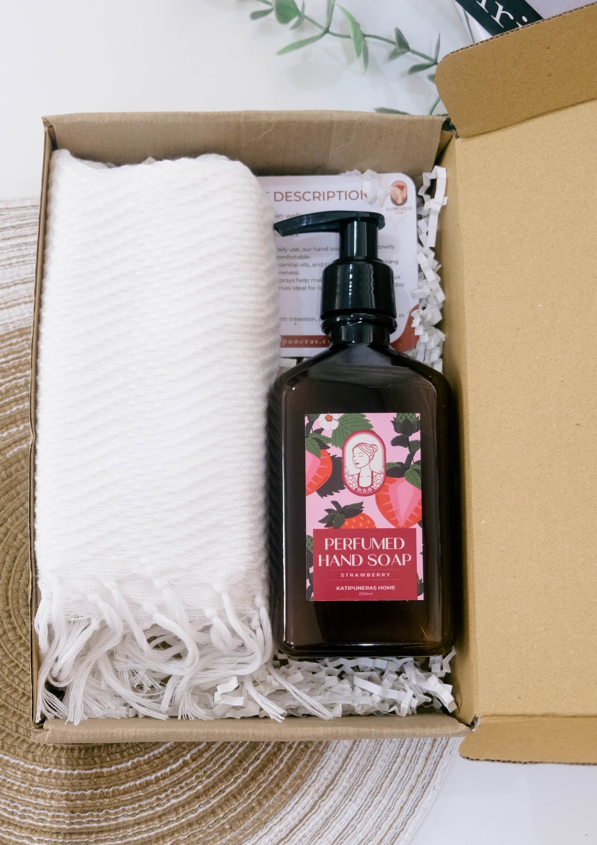 A dark amber bottle of strawberry-infused hand soap with a black pump, nestled in white paper shred inside a brown cardboard gift box next to a white textured towel. The soap label features pink strawberries and the text 'PERFUMED HAND SOAP STRAWBERRY'.