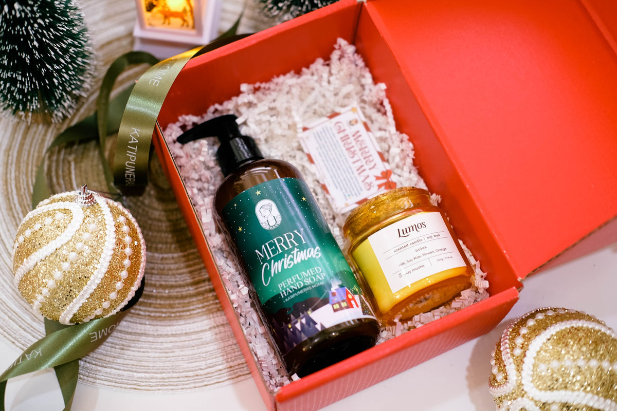 A red gift box filled with holiday-themed products, including a dark bottle labeled 'Merry Christmas' and a gold-labeled candle, surrounded by festive ornaments and ribbons on a tabletop.