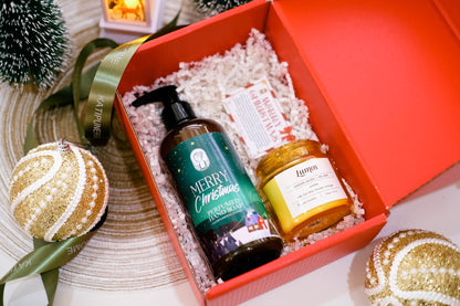 A red gift box filled with holiday-themed products, including a dark bottle labeled 'Merry Christmas' and a gold-labeled candle, surrounded by festive ornaments and ribbons on a tabletop.
