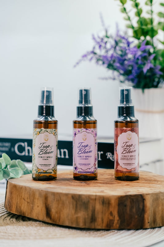 Three amber spray bottles labeled 'Trap + Bloom' in lemon, lavender, and Wood scents, displayed on a round wooden slice with a blurred background of flowers and books.