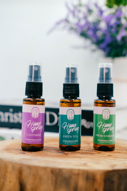 Three small amber home spray bottles with black spray tops are lined up on a wooden surface. The labels read Home Spray Lavender, Home Spray Green Tea, and Home Spray Fresh Bamboo, with a blurred background showing purple flowers and a white wall.