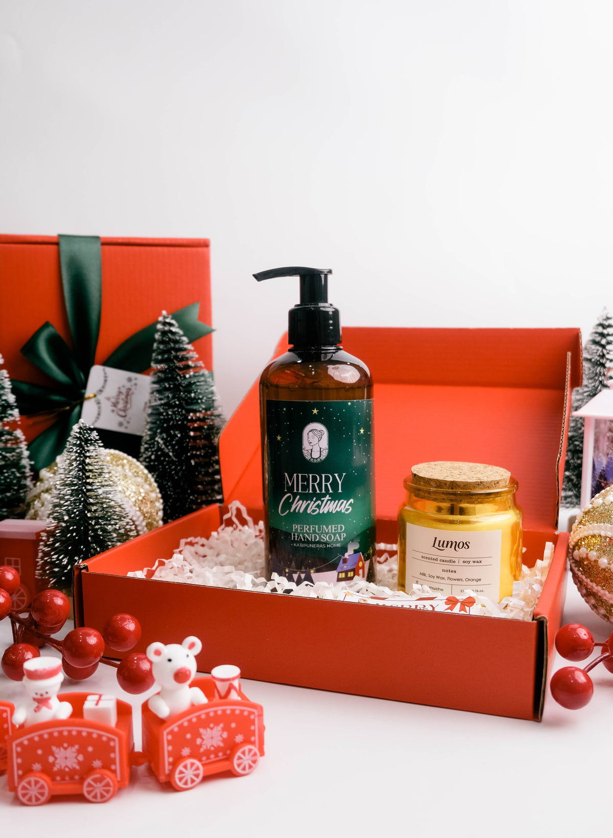 A festive Christmas gift set in an open red box, featuring a dark amber pump bottle labeled 'Merry Christmas' and a small gold candle jar, surrounded by holiday decorations including miniature trees, red ornaments, and a red gift box in the background.